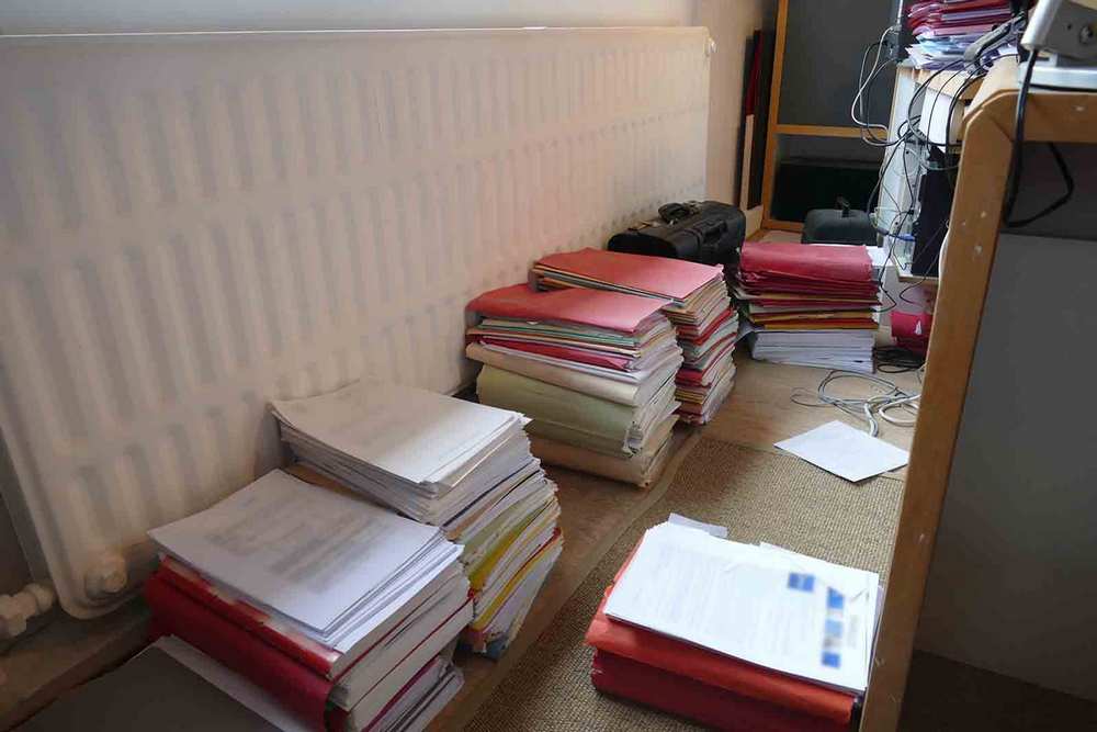 Plusieurs piles de dossiers et documents dans le bureau d’un avocat qui fait du pro deo à Namur. Une mallette d’ordinateur est également au sol.