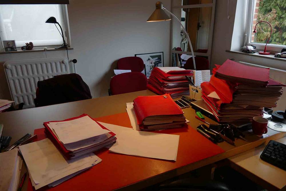Bureau d’un avocat de Namur qui fait du pro deo. Nombreuses piles de dossiers rouges et orange, sous-main rouge, lampe de bureau allumée.